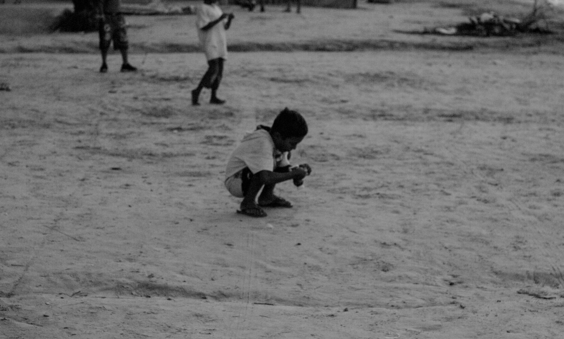Criança agachada brincando na areia, ao fundo outras duas crianças